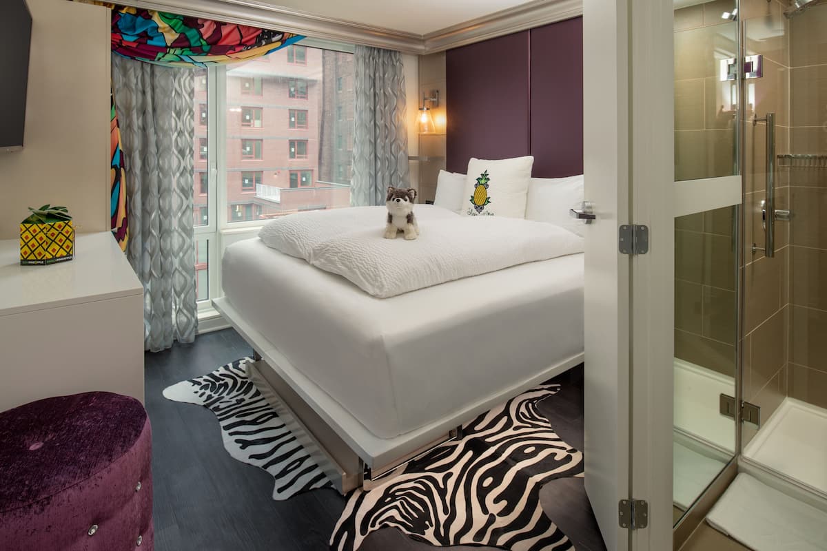 A hotel room with a large bed, a zebra rug, a plush purple ottoman, a pineapple decoration, and a view of buildings through the window.