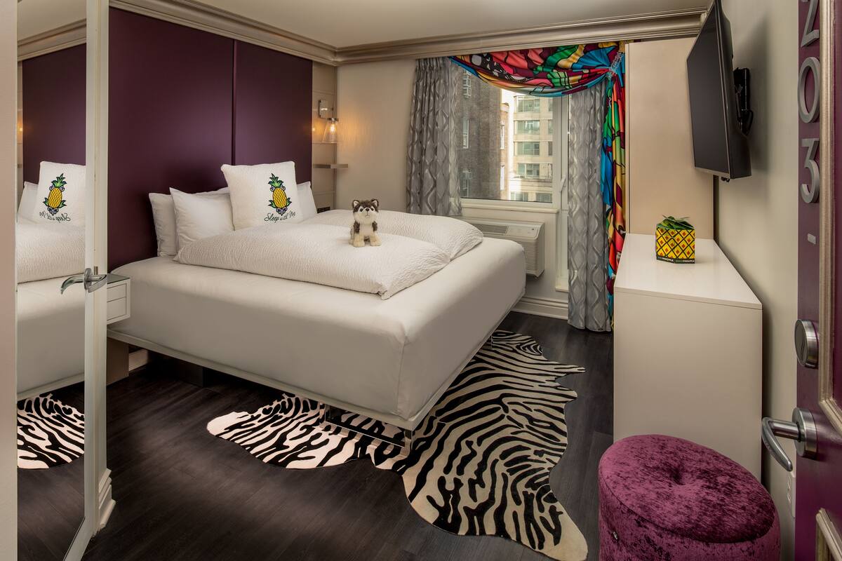 A modern hotel room with a large bed, a zebra-patterned rug, a small purple ottoman, and a television on the wall.