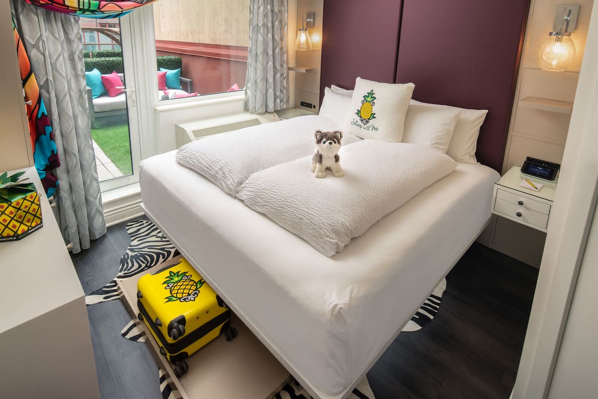 A hotel room with a bed, a small table, a nightstand, a suitcase, and a view of the outside.