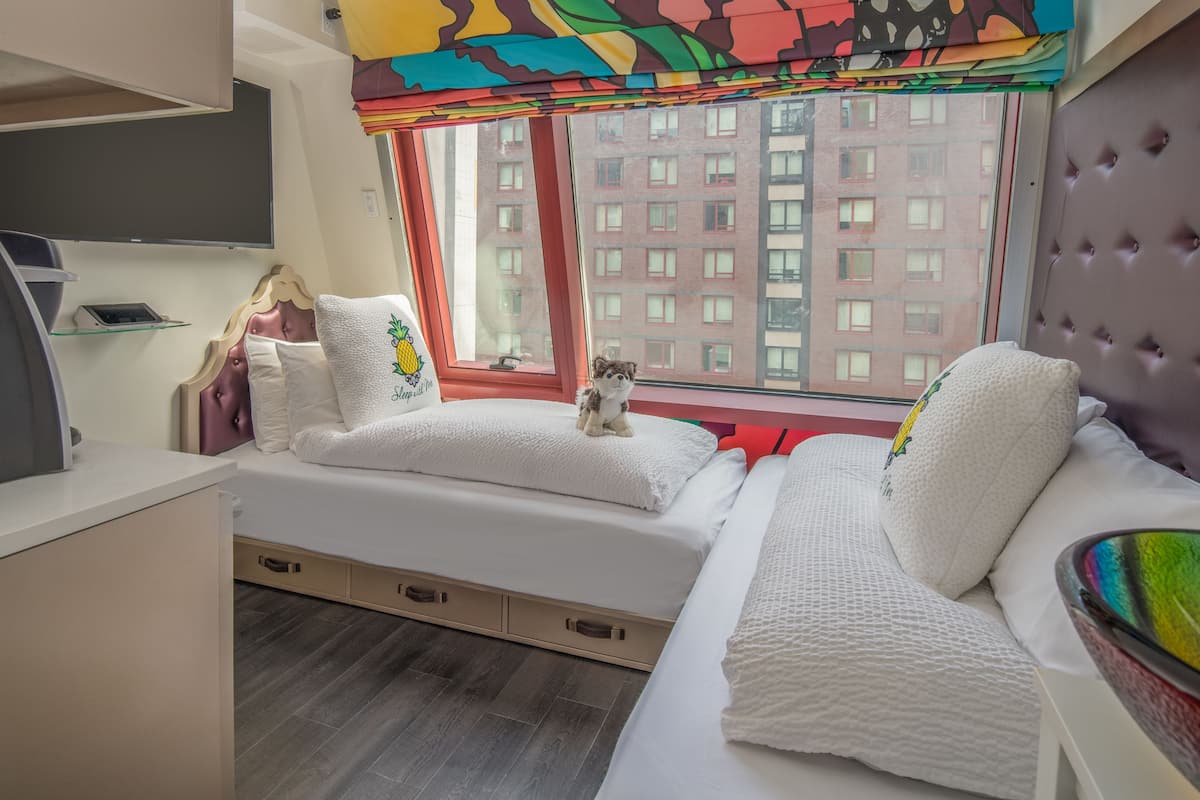 A modern hotel room with two beds, a window with a view of a red building, and a colorful curtain.