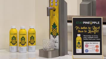 Dispensador de agua con tres botellas de agua Staypineapple, un vaso y un cartel que promueve el llenado de botellas en lugar del descarte.