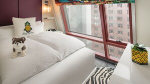 Quarto de hotel com uma cama, um brinquedo de pelúcia, uma almofada com padrão de abacaxi, uma cortina de janela colorida e uma caixa com padrão de abacaxi no criado-mudo.