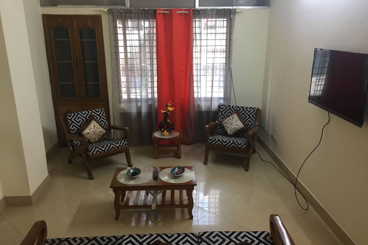 A living room with two patterned armchairs, a coffee table, a TV mounted on the wall, and a wooden cabinet.