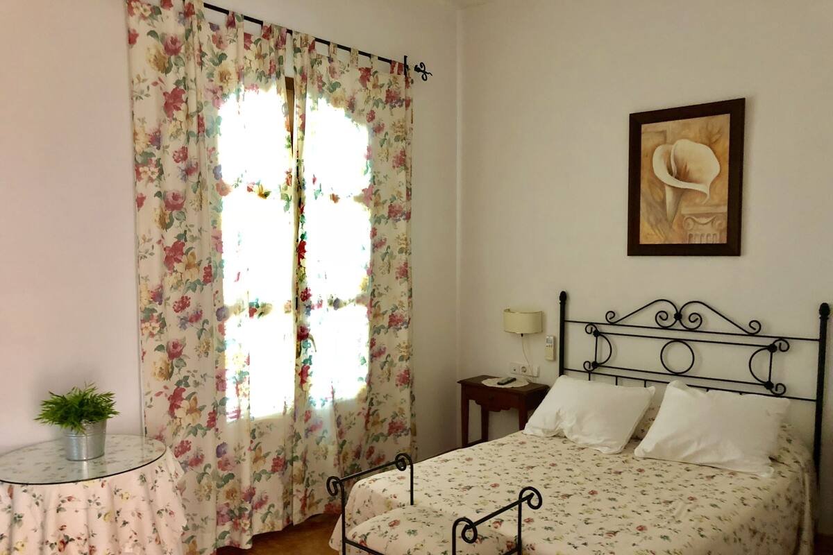 Une chambre à coucher avec une couvre-lit à motifs floraux, un cadre de lit en métal, une table de chevet avec une lampe et une œuvre d’art encadrée accrochée au mur.