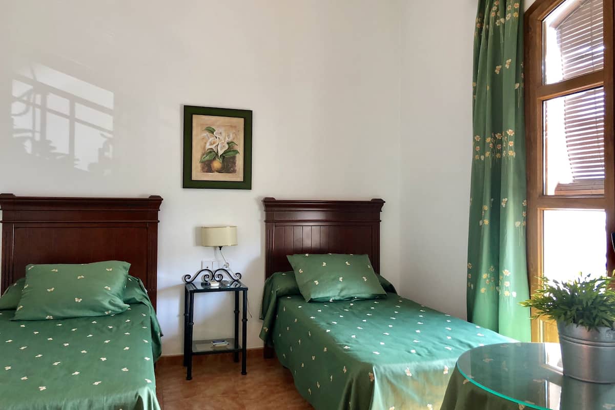 Une chambre avec deux lits, une petite table avec une lampe, un tableau encadré au mur et une fenêtre avec des rideaux.