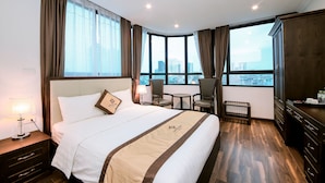 A hotel room with a large bed, a desk, two chairs, a view of the city, and a wardrobe. - Louisland Hotel (Hanoi)
