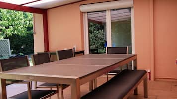 Een overdekt terras met een lange tafel, banken, grote ramen en uitzicht op groen.