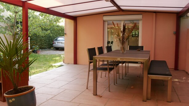 A covered patio with a table and chairs, a potted plant, and a view of a car and greenery outside. - Villa Seignemartin (Lyon)