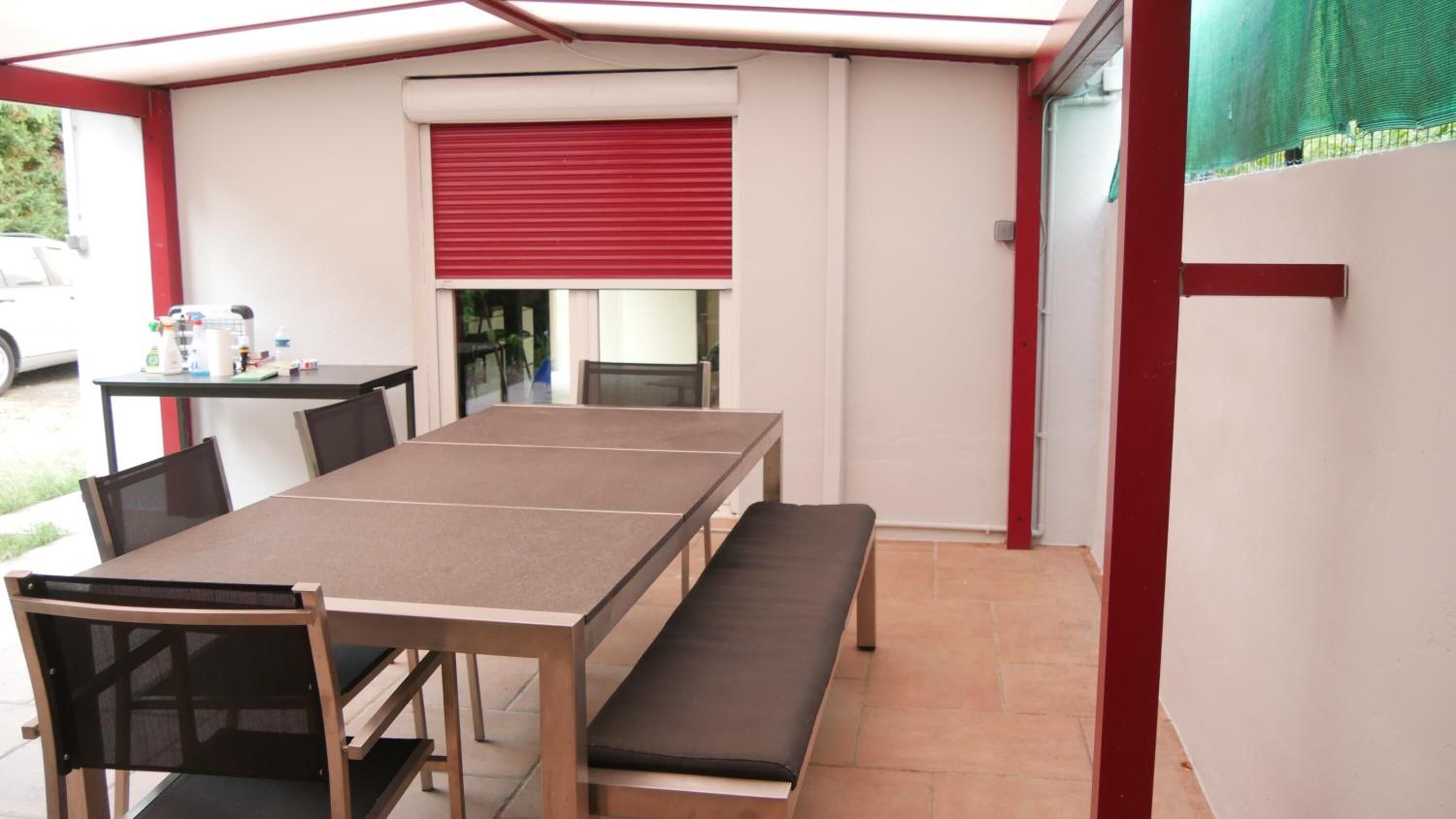 Un patio techado con una mesa, sillas y un banco, además de una puerta con contraventanas rojas.