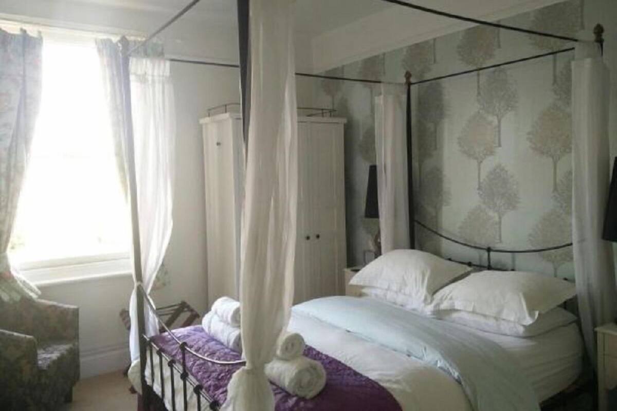 A four-poster canopy bed with white bedding, a wooden wardrobe, and a patterned wallpaper.