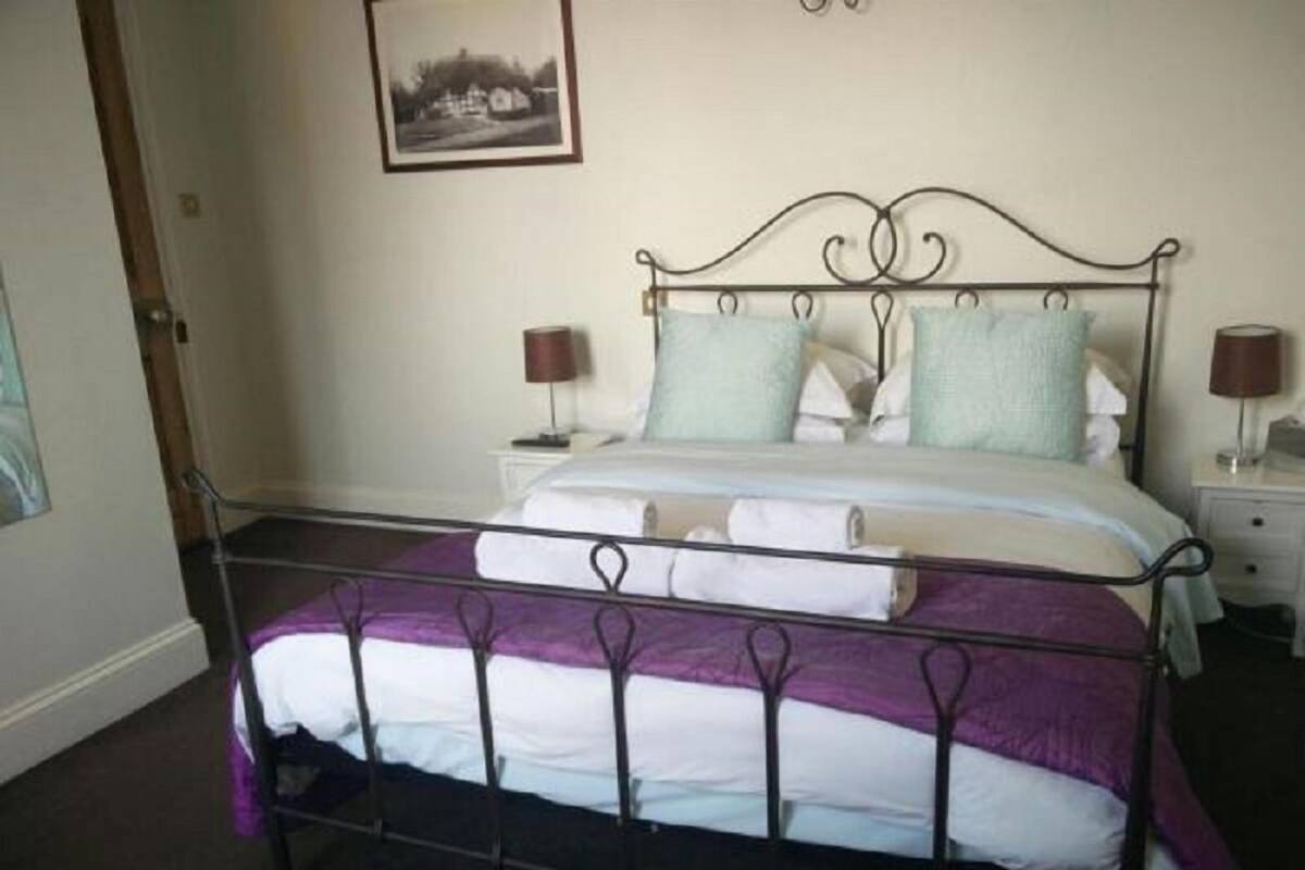 A neatly made bed with a metal frame, white bedding, and a purple throw. There are two bedside tables with lamps, and a framed picture on the wall.