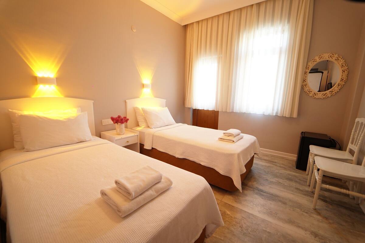 A hotel room with two beds, a nightstand with a lamp, a chair, and a large window with curtains.