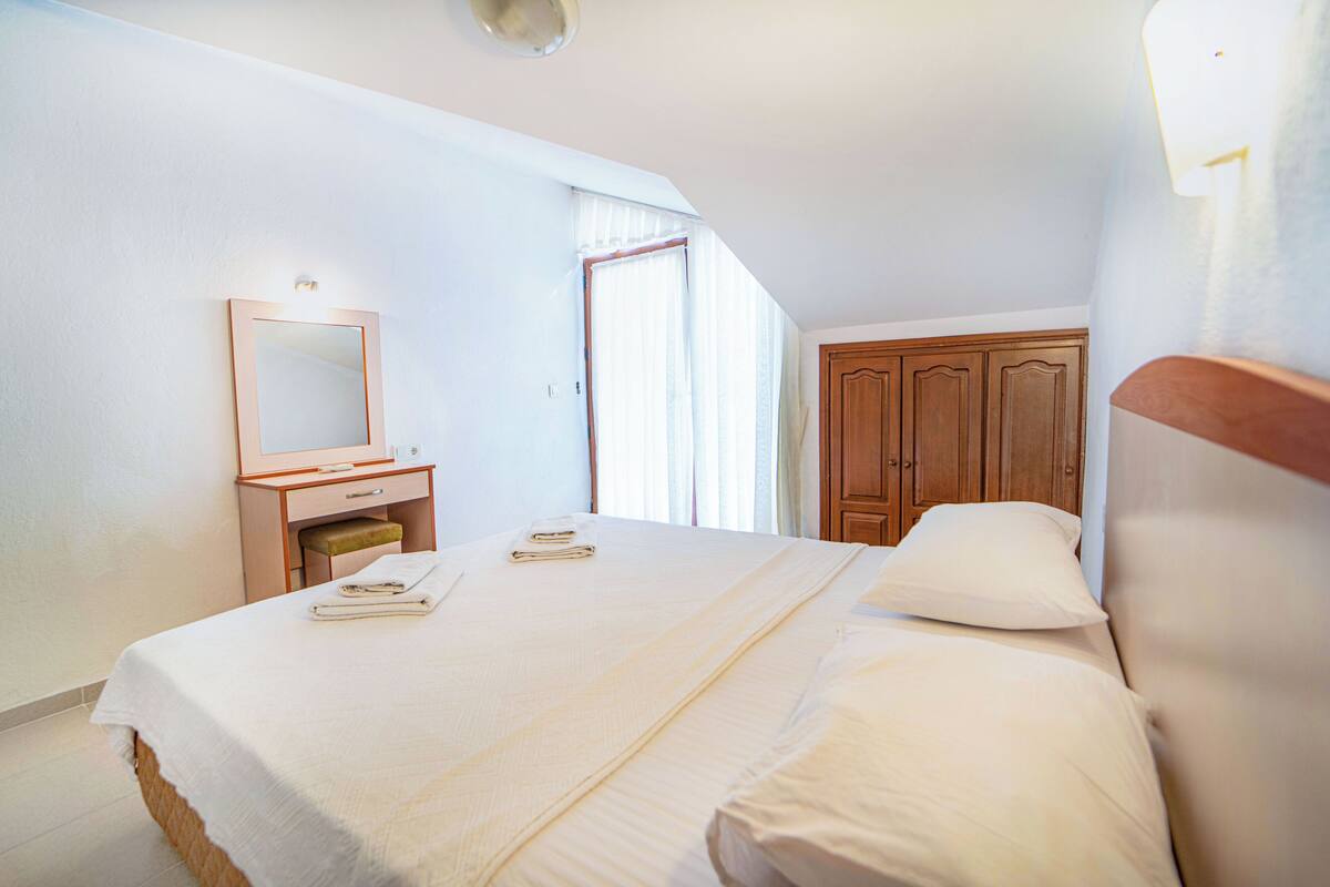 A bedroom with a bed, a wooden wardrobe, a bedside table with a mirror, and a window with curtains.
