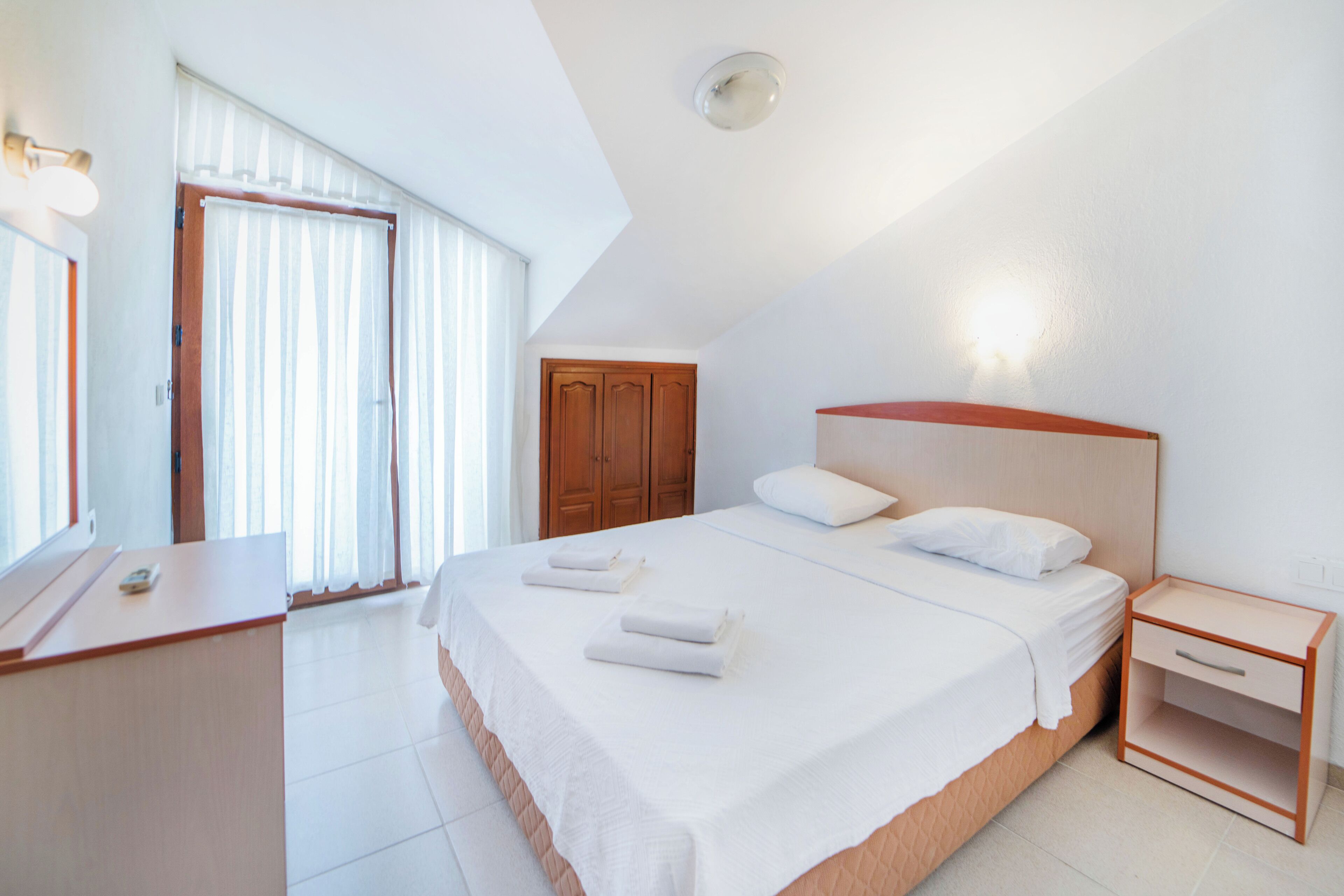 Habitación de hotel con una cama grande, un armario de madera, una mesita de noche y una ventana con cortinas.