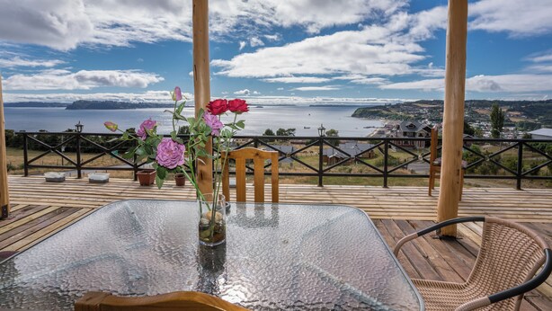 Una mesa de vidrio con un florero, una silla de mimbre y una vista de un lago y terreno lejano.