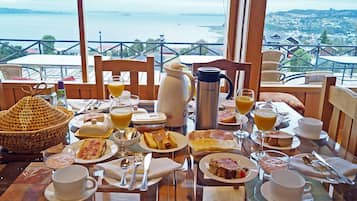 Una mesa de desayuno preparada con diferentes platos, una cafetera y vistas a un paisaje costero.