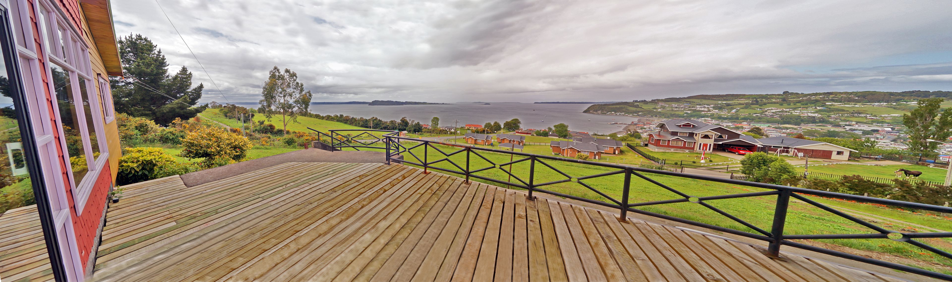 A wooden deck with a metal railing overlooking a coastal town with houses and a body of water.
