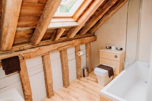 A small, rustic bathroom with wooden beams, a skylight, a toilet, a sink, and a bathtub. - Creeksea Place Barns (Burnham-on-Crouch)