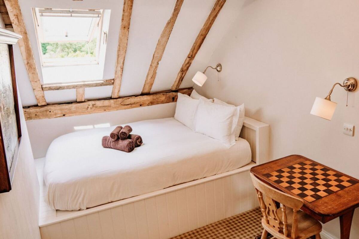 A cozy bedroom with a wooden ceiling, a bed with white linens, a small table with a chessboard, and a window with a view of greenery.