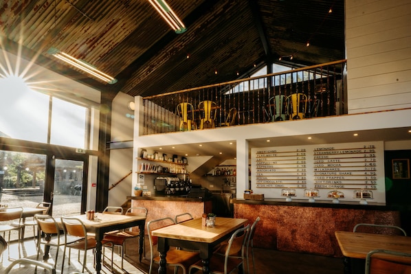 Un restaurante con decoración rústica, que presenta vigas de madera, sillas de metal y una pizarra con varios refrescos y platos.
