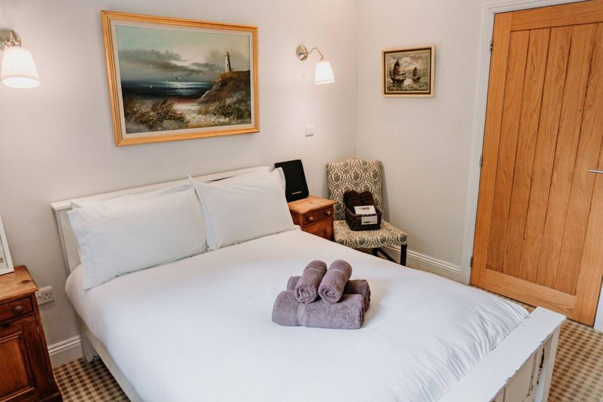 A neatly made bed with white linens, a set of towels, a chair with a patterned cushion, and a wooden door.