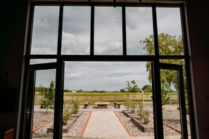An open window with a view of a paved outdoor area with benches and a picnic table, surrounded by a wooden fence and trees. - Creeksea Place Barns (Burnham-on-Crouch)