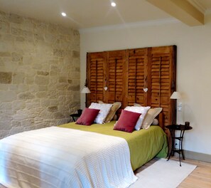 A bedroom with a stone wall, wooden headboard, two bedside tables with lamps, a bed with a green bedspread and white bedding, and a small round table with a bowl and candle. - Demeure d'Hôtes L'Hermitage (Aix-les-Bains)