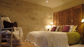 Ein Schlafzimmer mit Steinwand, Holzläden, einem Bett mit weißer und grüner Bettwäsche sowie einem Sessel mit weißer Decke.