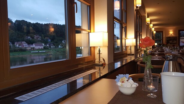 Un restaurante con vistas a un lago y edificios, una rosa en un florero y una vela sobre la mesa.