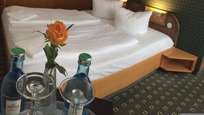 Una cama bien hecha con sábanas blancas, una rosa naranja y dos botellas de licor sobre una mesita de noche.