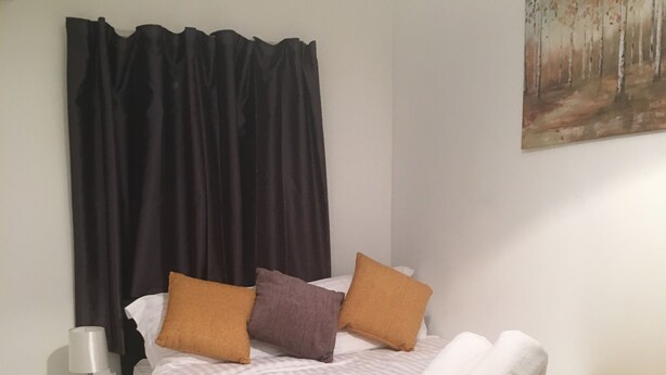 A neatly made bed with a white comforter, a mustard-colored throw blanket, and two pillows in brown and purple. There is a white bedside table with a lamp and a small drawer. The room has a wooden floor and a painting of a forest on the wall. - Kingswood Apartment (London)