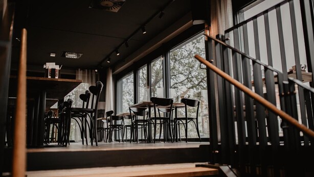 Vista interior de un restaurante con mesas y sillas dispuestas cerca de amplias ventanas, una escalera con barandilla metálica y un techo con iluminación empotrada.