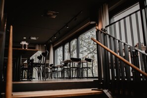 Vista interior de un restaurante con mesas y sillas dispuestas cerca de amplias ventanas, una escalera con barandilla metálica y un techo con iluminación empotrada.