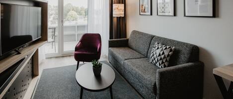 A modern living room with a grey sofa, a red armchair, a small round table with a plant, and a TV on a wooden stand.