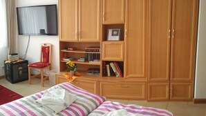 A hotel room with a large wooden wardrobe, a flat-screen TV mounted on the wall, a chair, a bed with striped bedding, and a small table with a vase of flowers. - Privatunterkunft AARON (Magdeburg)