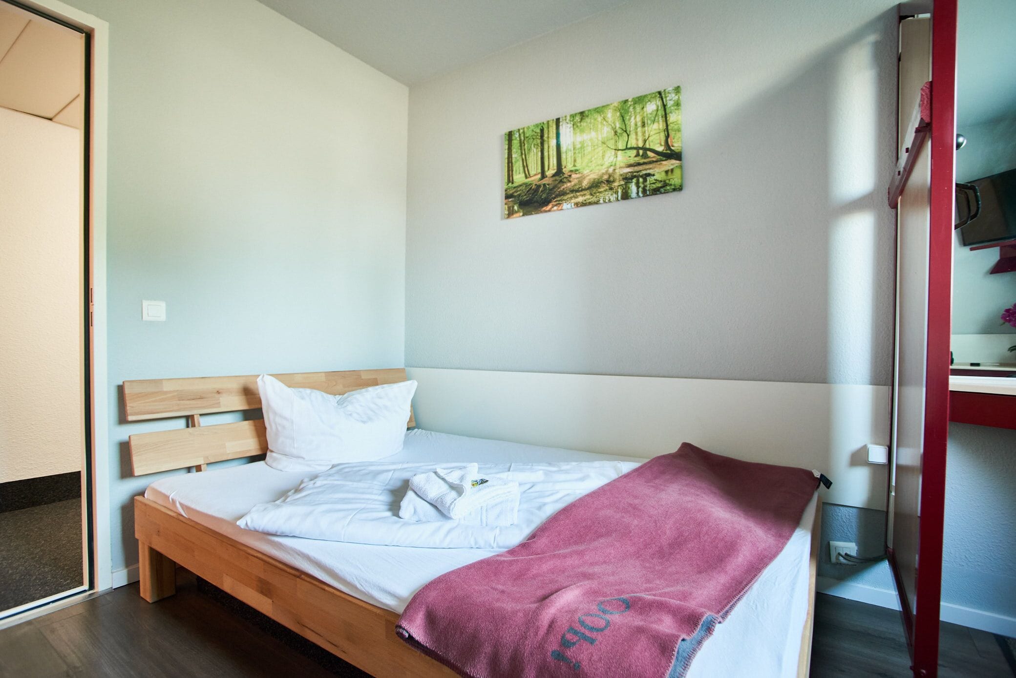 Una cama individual con cabecera de madera, ropa de cama blanca y una manta roja. En la pared sobre la cama hay un cuadro de un bosque.