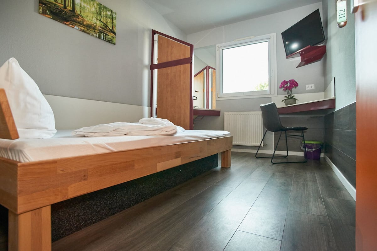 A bedroom with a wooden bed, a desk with a chair, a TV mounted on the wall, and a window.