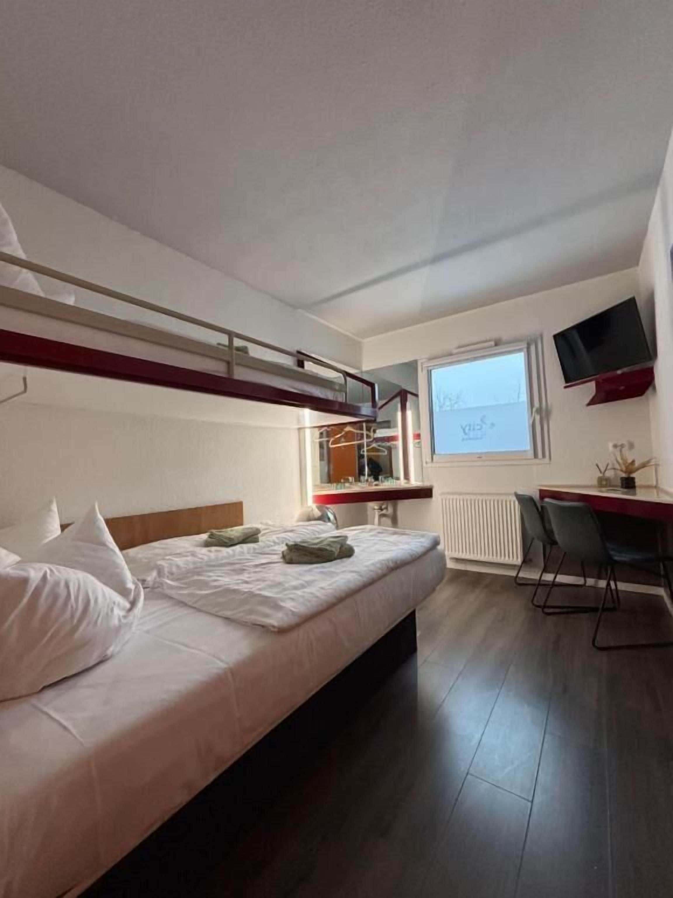 A hotel room with a bunk bed, a desk, a chair, a TV, and a window with a view.