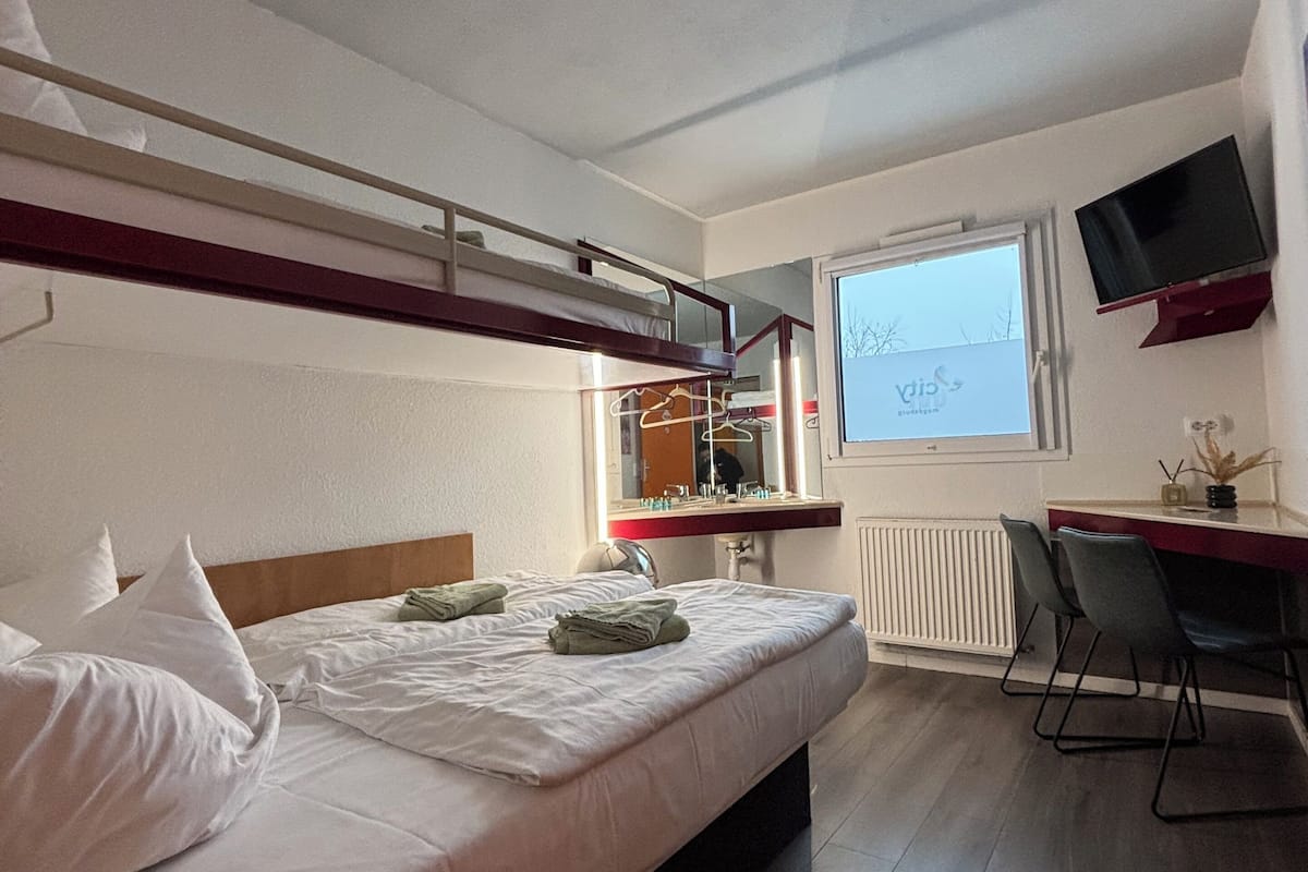 A hotel room with a bunk bed, a desk with a chair, a television, and a window with a view of the sky.