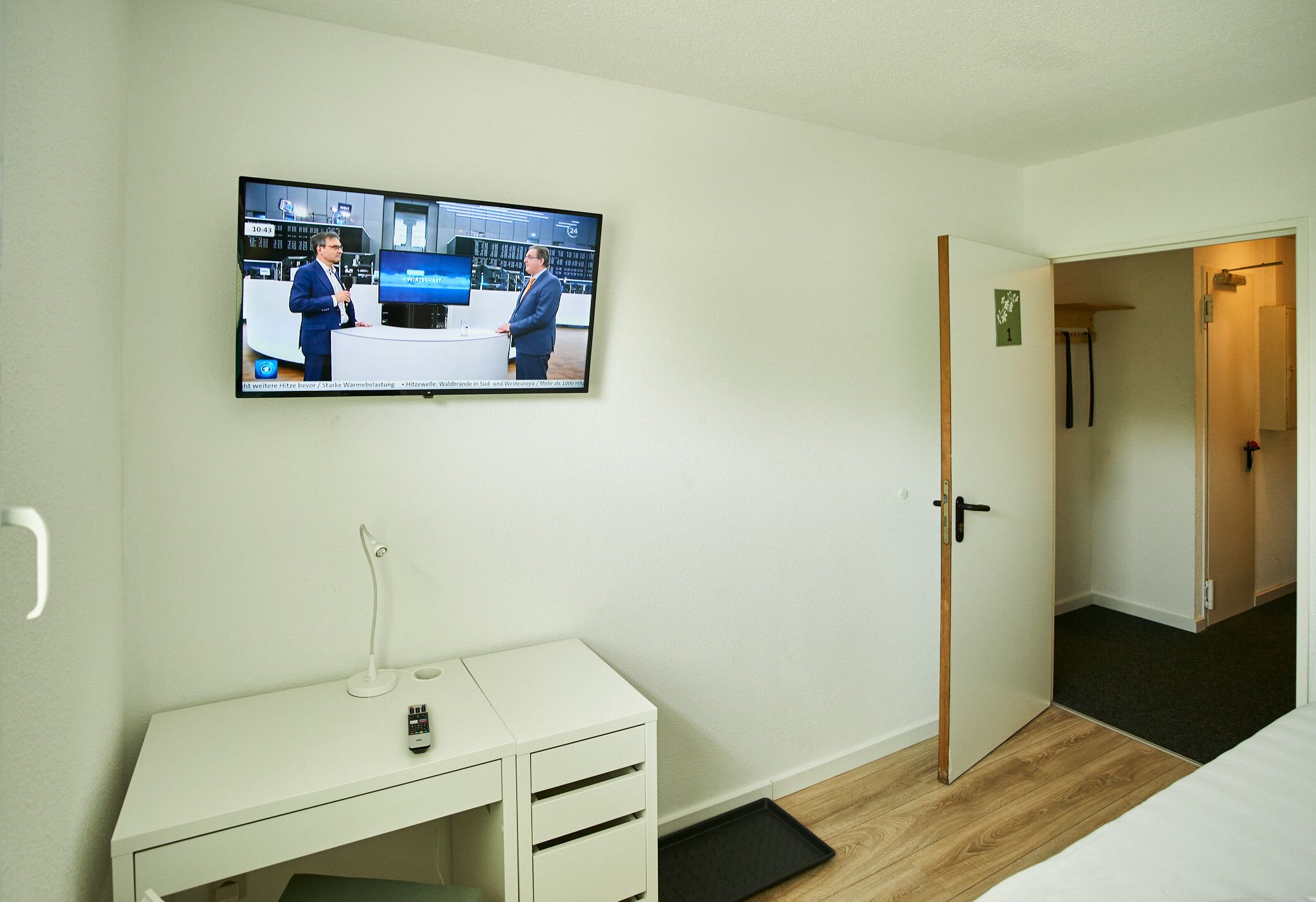 A hotel room with a TV mounted on the wall, a desk with a lamp, and a door leading to another room.