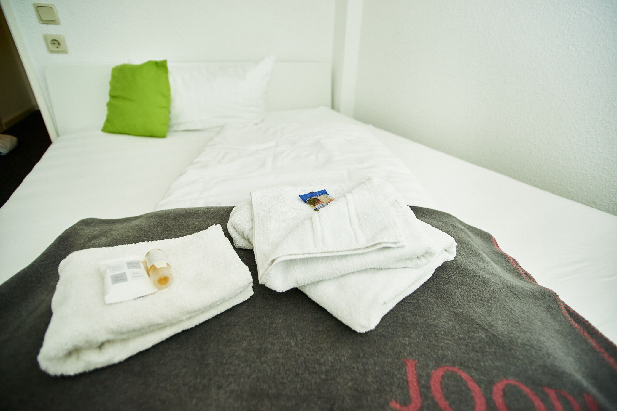 Una cama bien hecha con sábanas blancas, una almohada verde y una toalla con una botella encima.