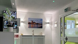 A reception area with a white reception desk, a mounted television displaying multiple screens, a painting of a cityscape, and a glass door entrance. - Hotel City Inn Magdeburg (Magdeburg)