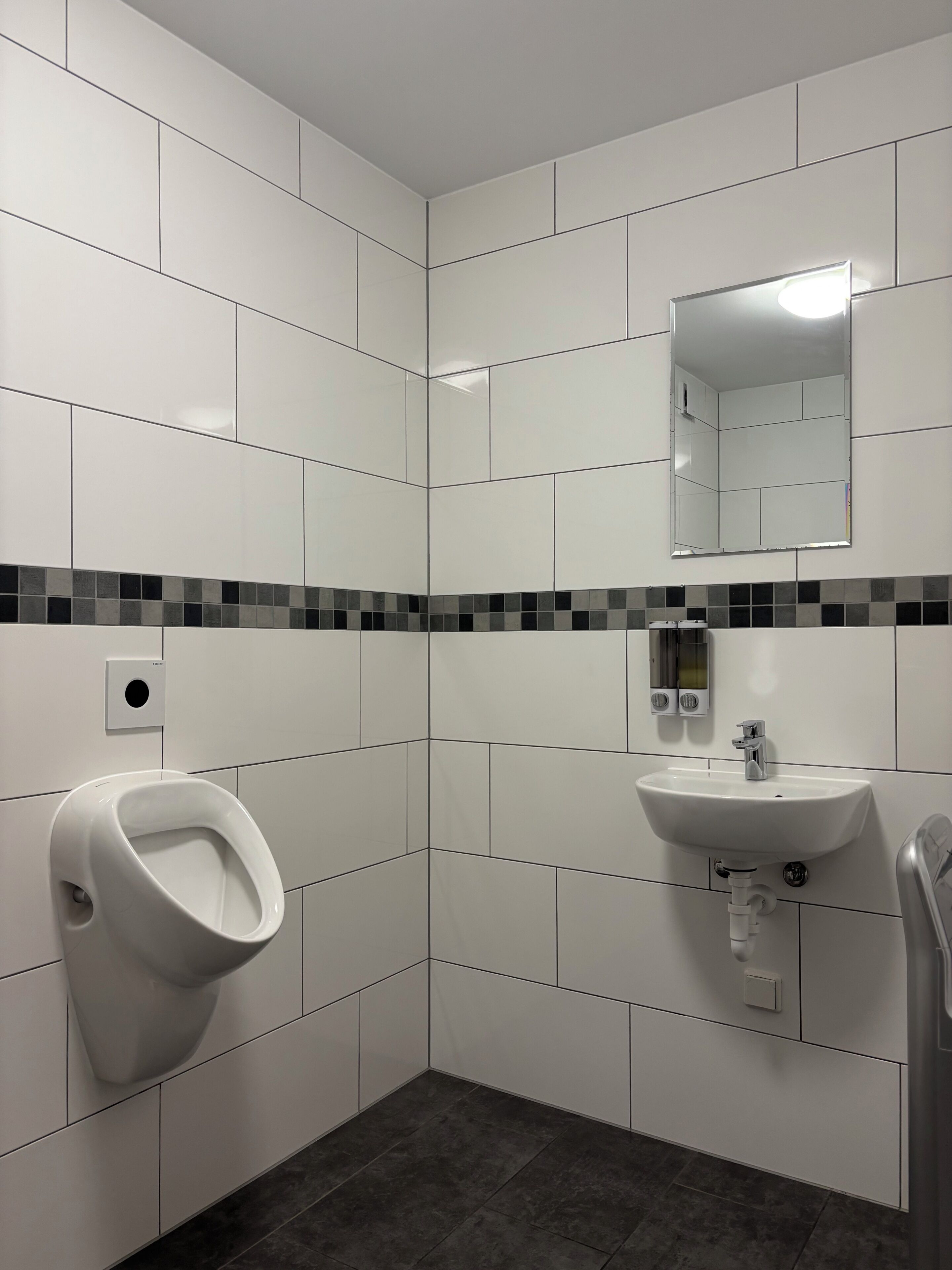 Un baño moderno con azulejos blancos, un orinal, un lavabo y un espejo.