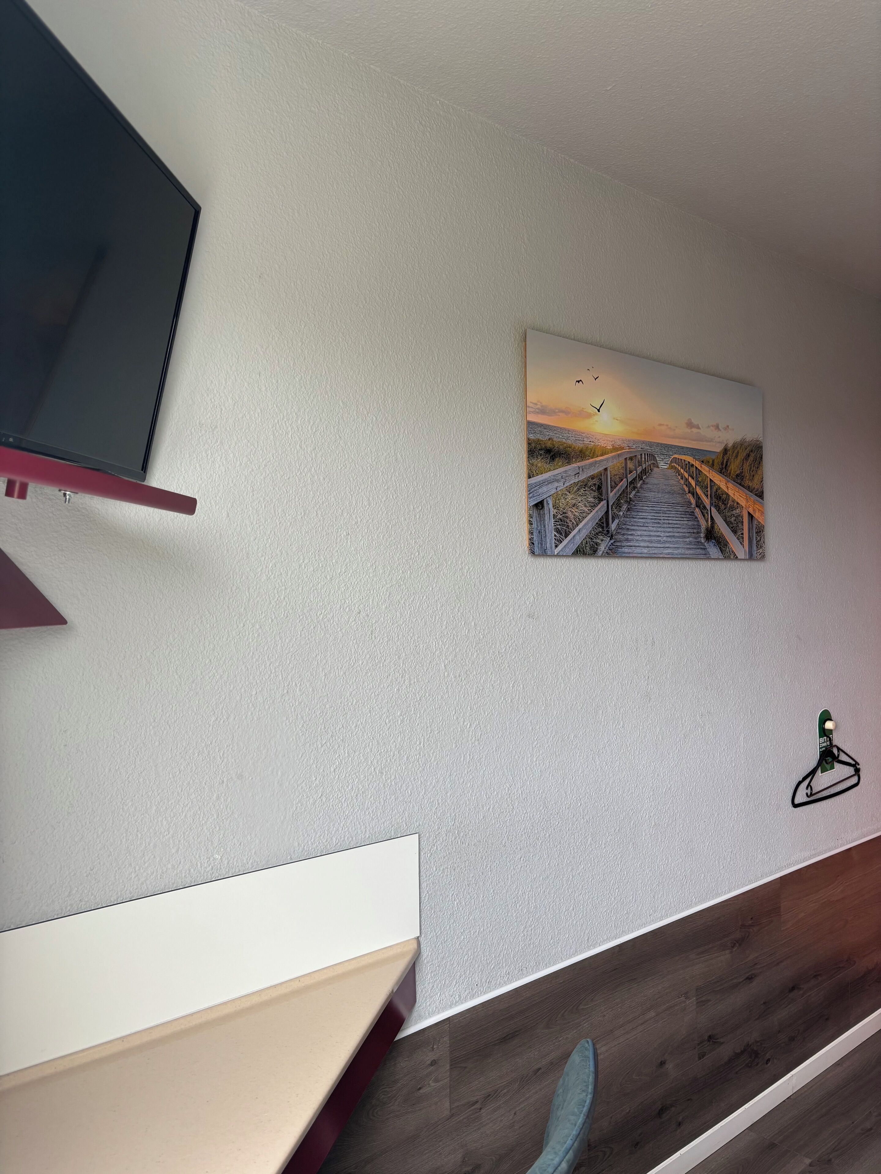 Ein Zimmer mit Fernseher, einem Sonnenuntergangsbild, einem kleinen grünen Haken und Holzboden.
