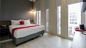 A modern hotel room with a large bed, a desk with a computer, and a view of the city through large windows. - RedDoorz Plus near Tebet (Jakarta)