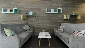 A modern living room with two grey sofas, a white coffee table, and three teal and yellow floating shelves against a wooden wall. - RedDoorz Plus near Tebet (Jakarta)
