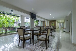 A spacious indoor area with multiple tables and chairs, a flat-screen TV mounted on a pillar, and large windows offering a view of greenery outside. - Sans Vibes Jasmine Suite TB Simatupang (Jakarta)