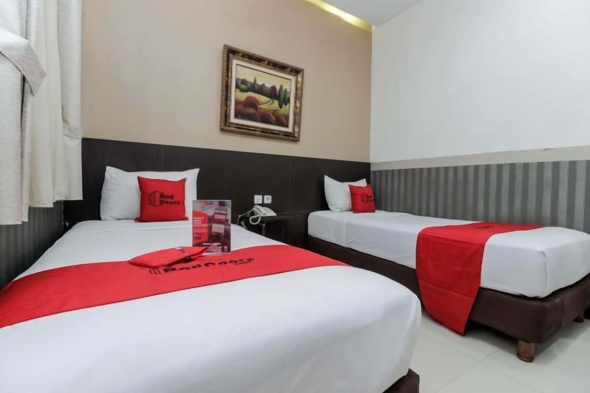 Habitación de hotel con dos camas, cada una con ropa blanca y detalles rojos, un cuadro de paisaje enmarcado en la pared y una manta roja sobre las camas.