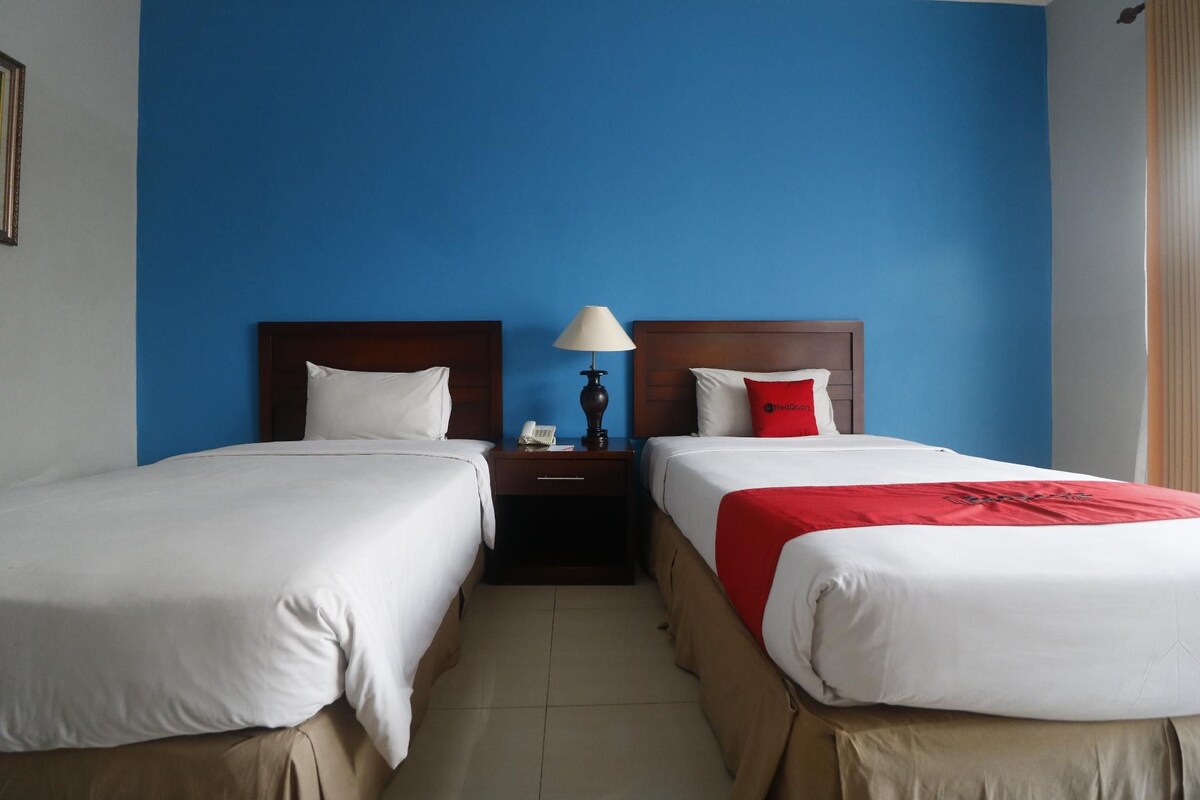 A hotel room with two single beds, a nightstand with a lamp, and a red throw blanket.