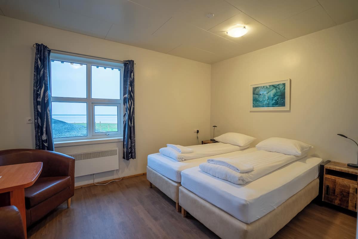 A room with two beds, a wooden table, a chair, a window with curtains, a painting on the wall, and a heater.